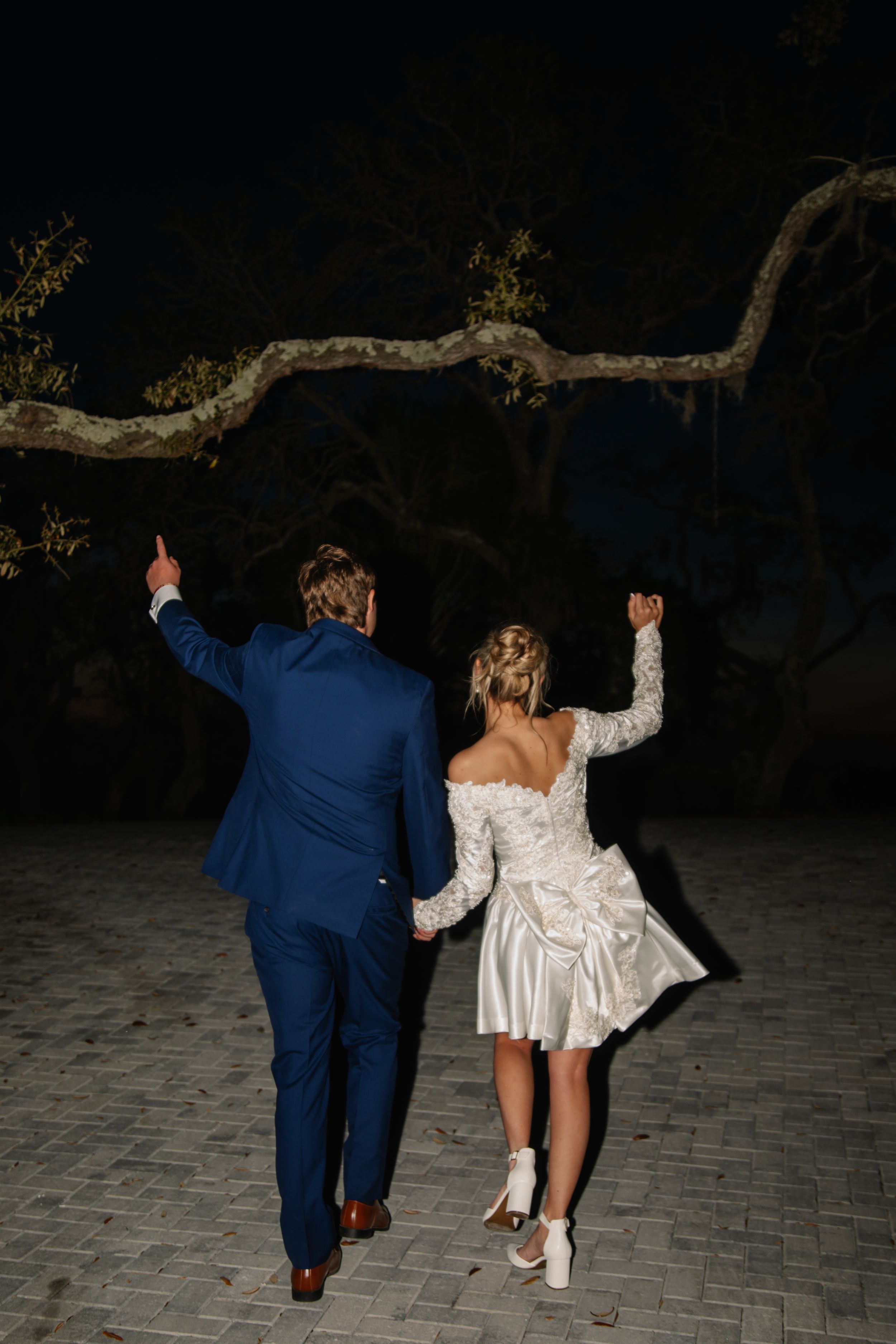A couple in wedding attire dances joyfully at night on a paved outdoor area beneath a twisted tree branch.