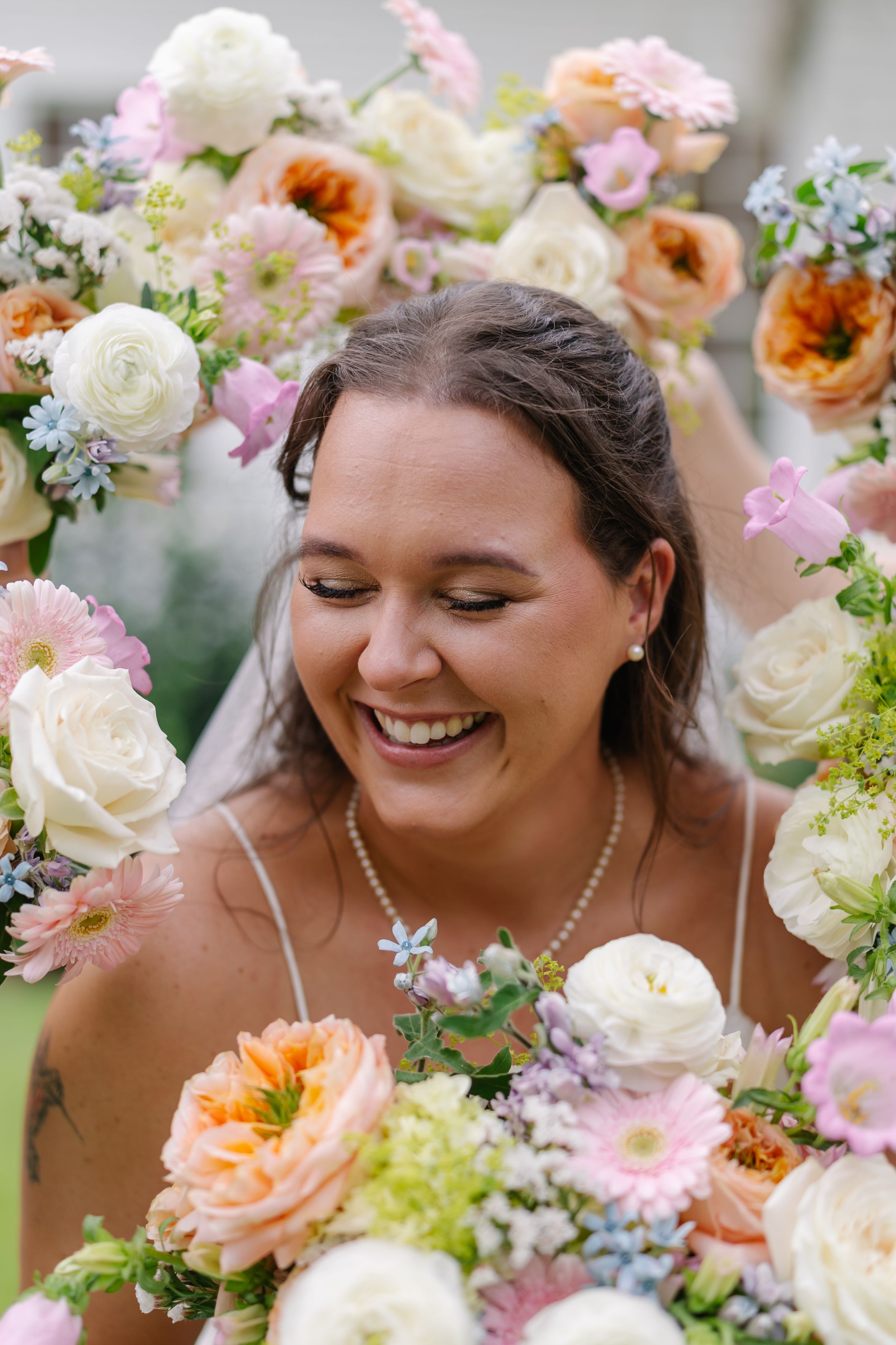 Smiling bride surrounded by colorful wedding bouquet of peach, white, and pink flowers including ranunculus and gerbera daisies.