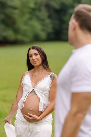 Pregnant woman in white dress holds baby bump outdoors, smiling at man in white shirt with green park background.