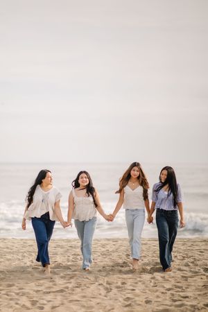 Four friends walking together holding hands and smiling on a sandy beach at sunset.