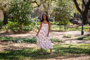 A flowing floral sundress is showcased in a sunlit garden path surrounded by trees and dappled light.