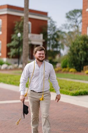 Smiling male college graduate walks on brick path holding cap and gold honor cords outside brick university building.