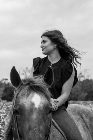 Black and white photo of a person with flowing hair riding horseback in a field on a cloudy day.