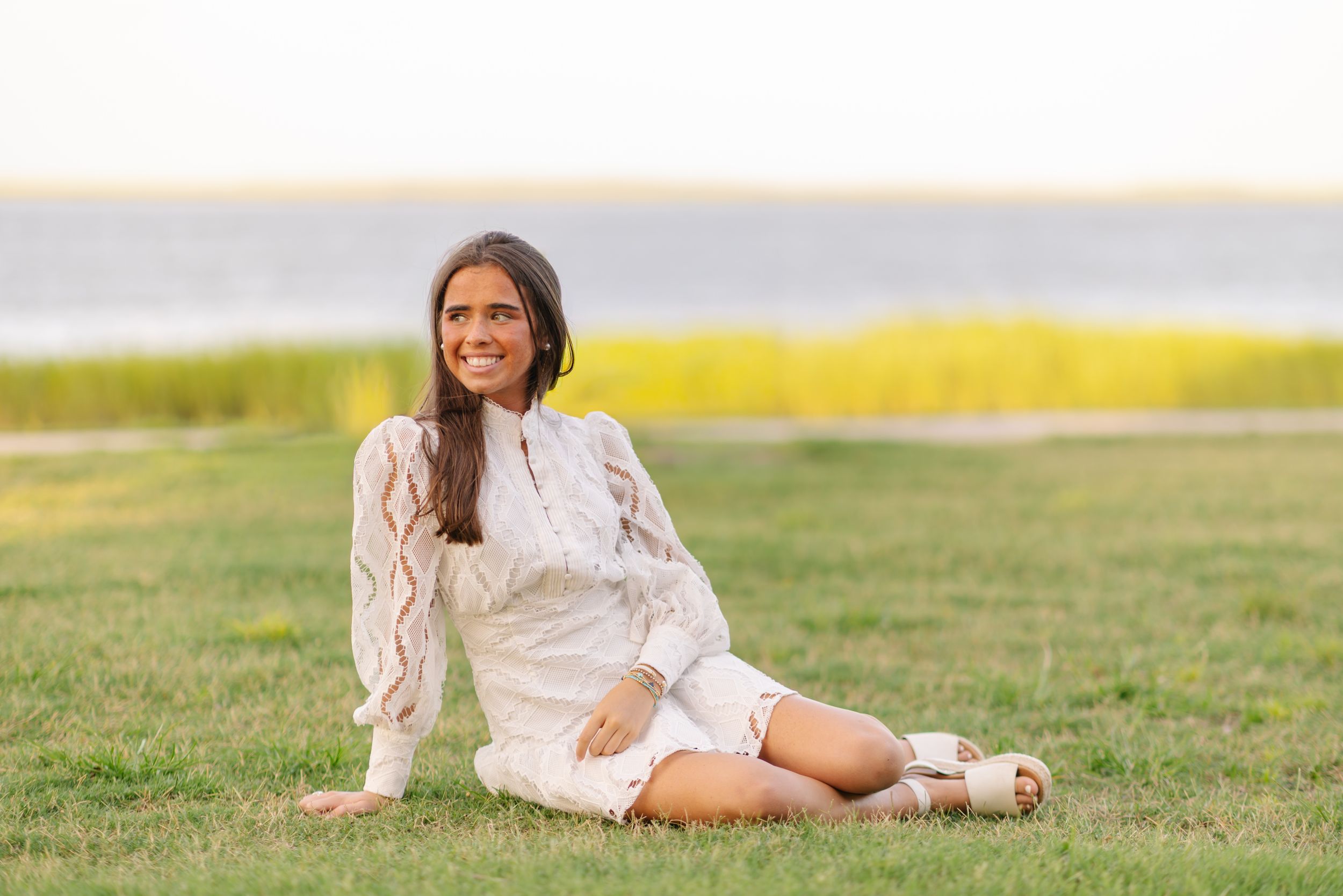 Young woman in white dress sitting on green grass by the water, smiling outdoors on a sunny day.