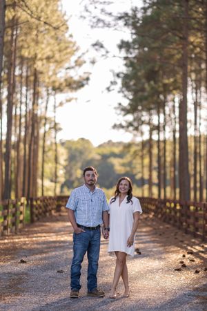 Couple holding hands on a tree-lined gravel path, man in plaid shirt and jeans, woman in white dress, golden hour light.