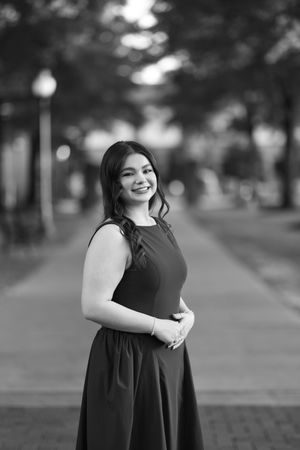 Black and white portrait of a smiling young woman in a sleeveless dress standing on a tree-lined brick pathway.