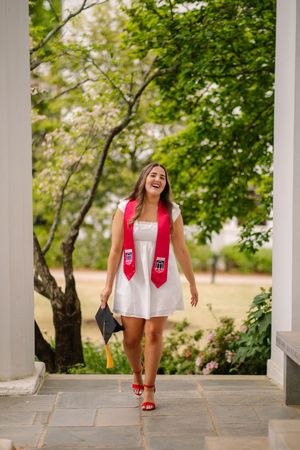 Graduate in white dress and red stoles walking confidently on stone pathway surrounded by lush green trees.