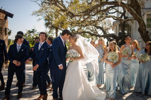 Wedding party in navy suits and light blue dresses standing outdoors under a large oak tree during sunny ceremony.
