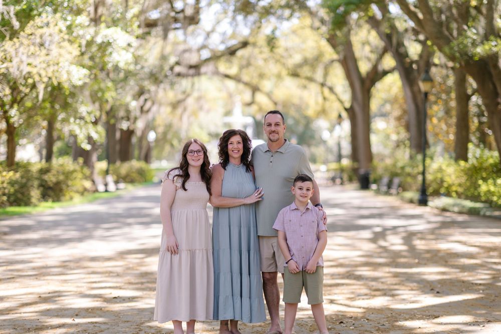 Family of four posing on a tree-lined path, wearing coordinated pastel outfits for an outdoor portrait session.