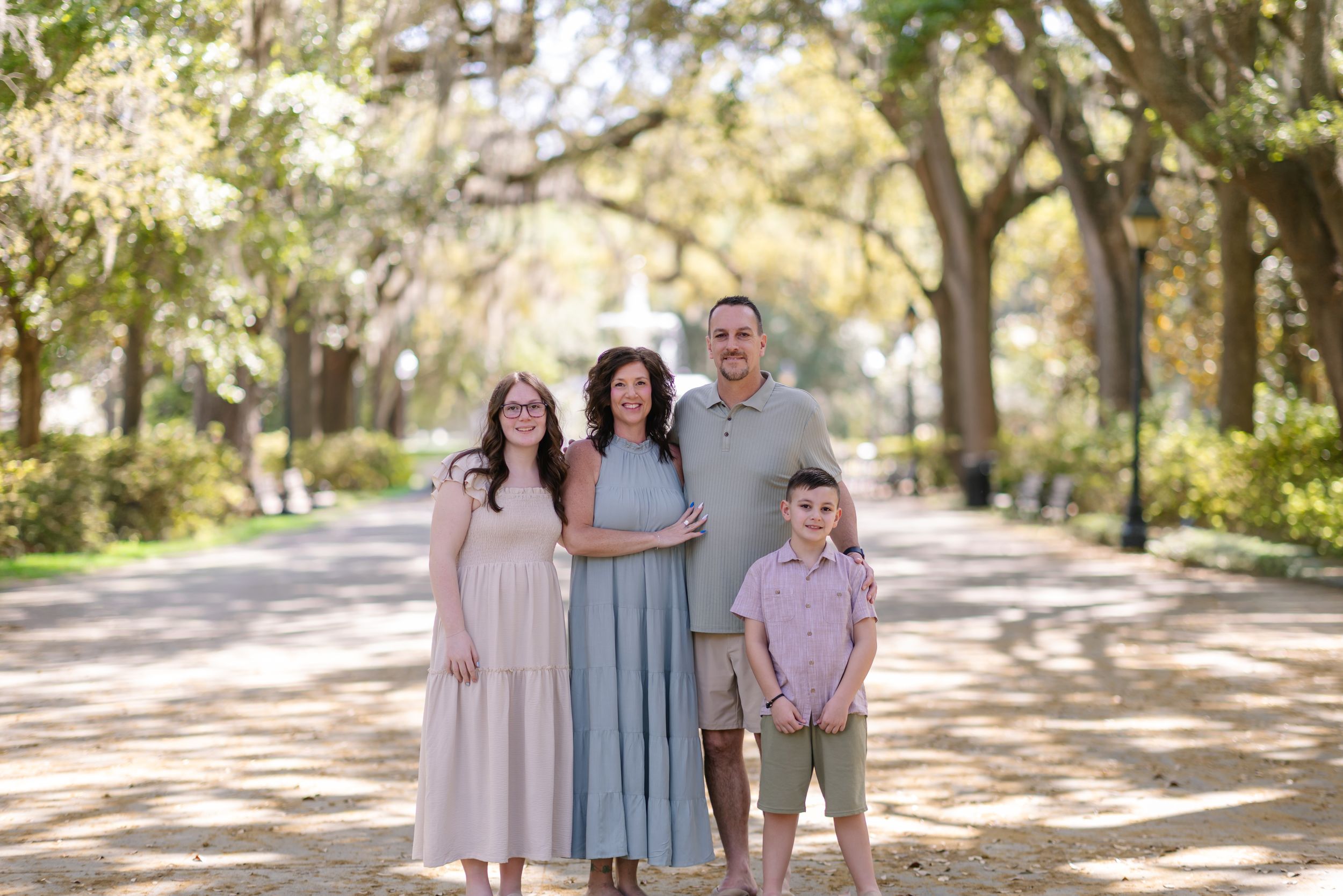 Family of four posing on a tree-lined path, wearing coordinated pastel outfits for an outdoor portrait session.
