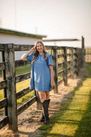 A denim dress and cowboy boots outfit styled in a rustic setting with wooden fencing at sunset.