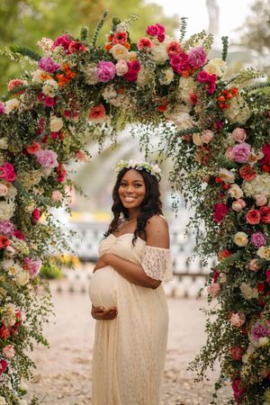Beautiful floral arch with pink and red roses frames an expectant mother in an off-shoulder cream lace dress and flower crown.