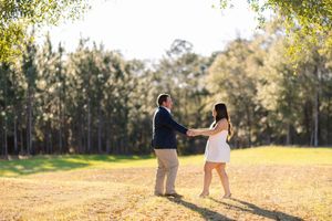 Couple dancing together in a sunlit field surrounded by tall green trees during a romantic outdoor engagement session.