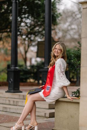 Smiling blonde graduate in white lace dress with red sash holds cap while sitting outdoors on stone ledge.