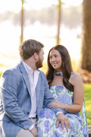 Couple sitting outdoors near a lake, woman in floral blue dress smiling at bearded man in light blue suit.