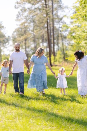 Family of five holding hands walking through a sunny green park, parents with two young children and a teen girl in white dress.
