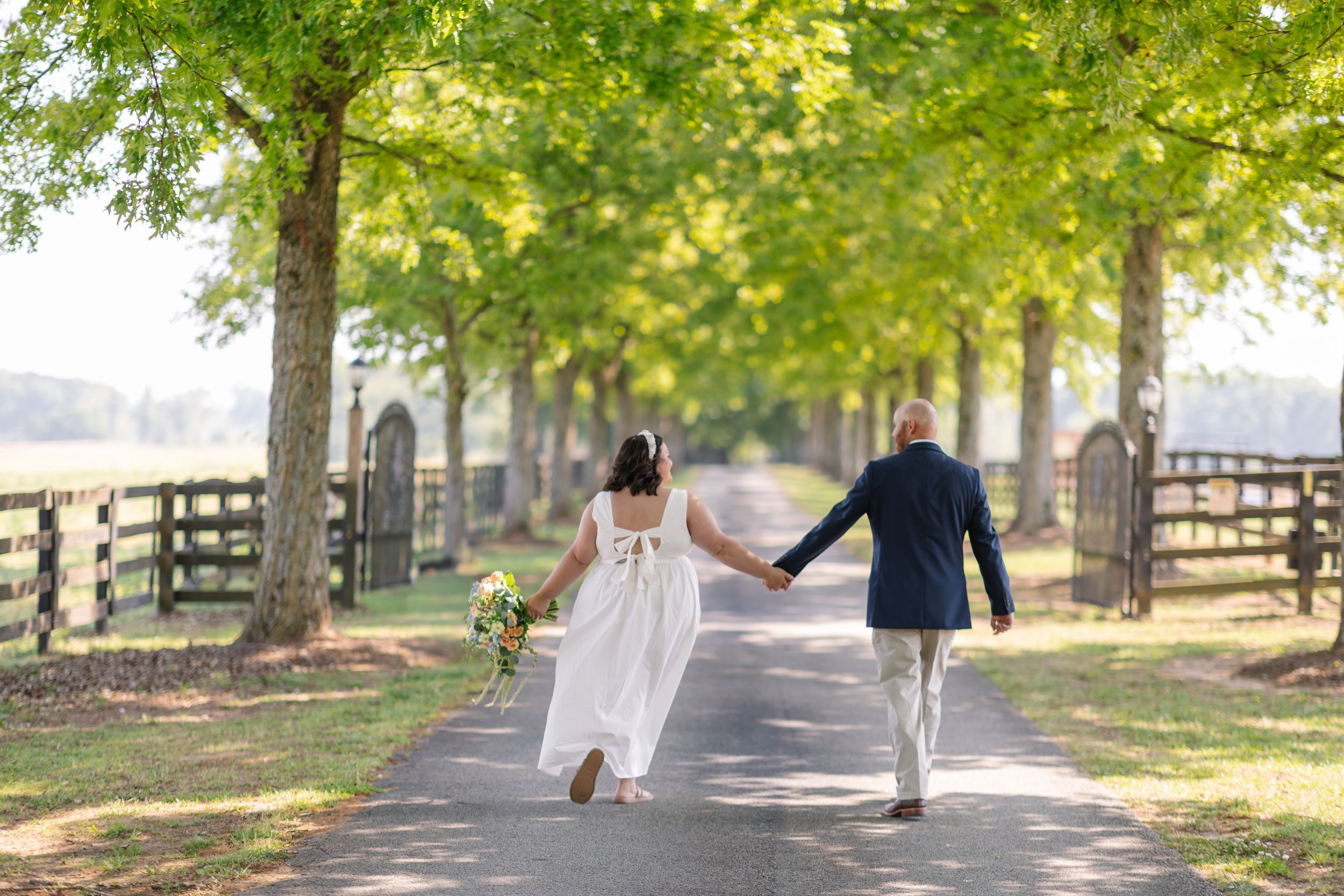 A bride and groom walk hand-in-hand down a tree-lined path at an outdoor wedding venue.