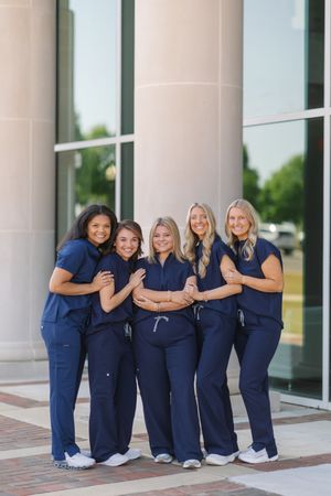 Five women in matching navy blue scrubs pose together smiling outside a modern building with large windows and columns.