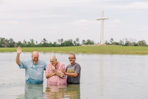 Three people performing a baptism ceremony in a lake with a large cross monument visible in the background.