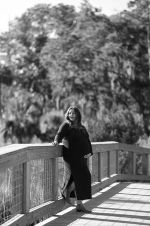 Black and white photo of a smiling pregnant woman in a dark dress leaning on a wooden bridge railing outdoors.