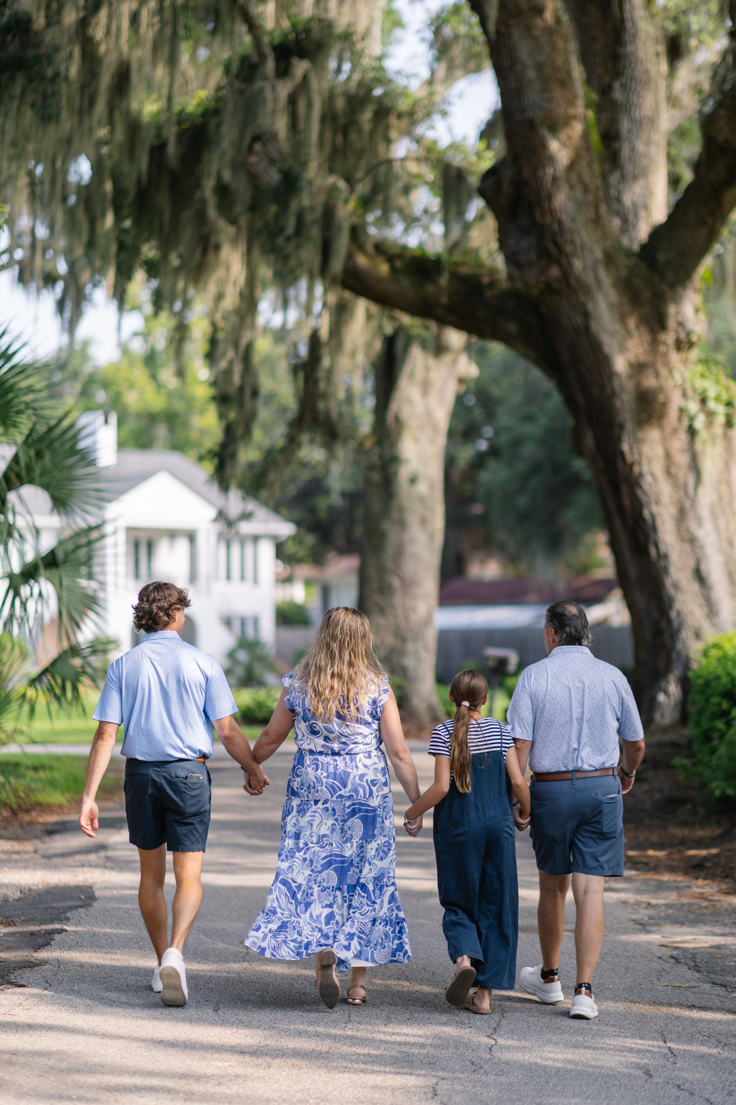 Family of four walking hand-in-hand down a tree-lined path draped with Spanish moss near a white home.