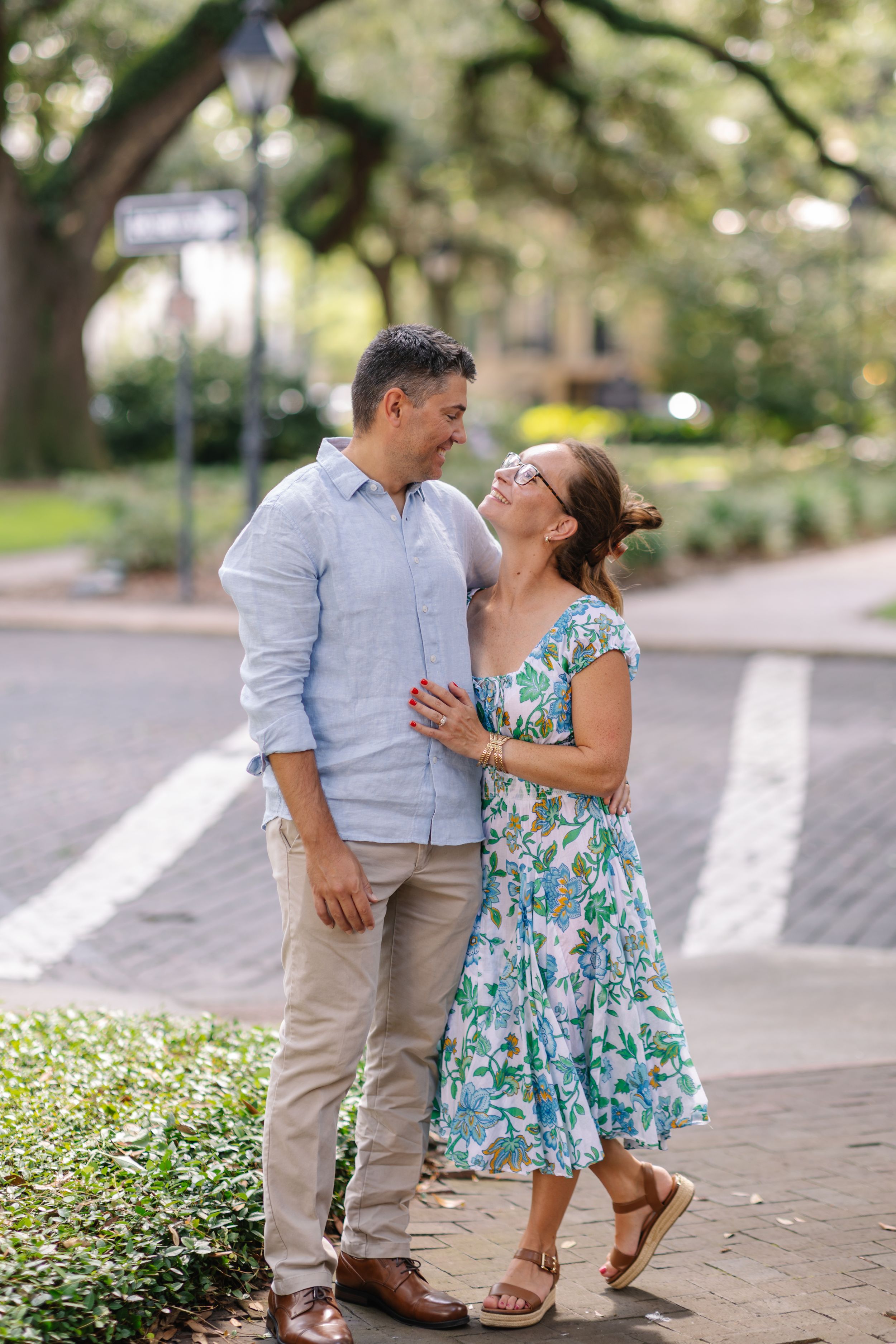 A couple shares a romantic moment on a Savannah, GA street, woman in floral dress, man in light blue shirt.