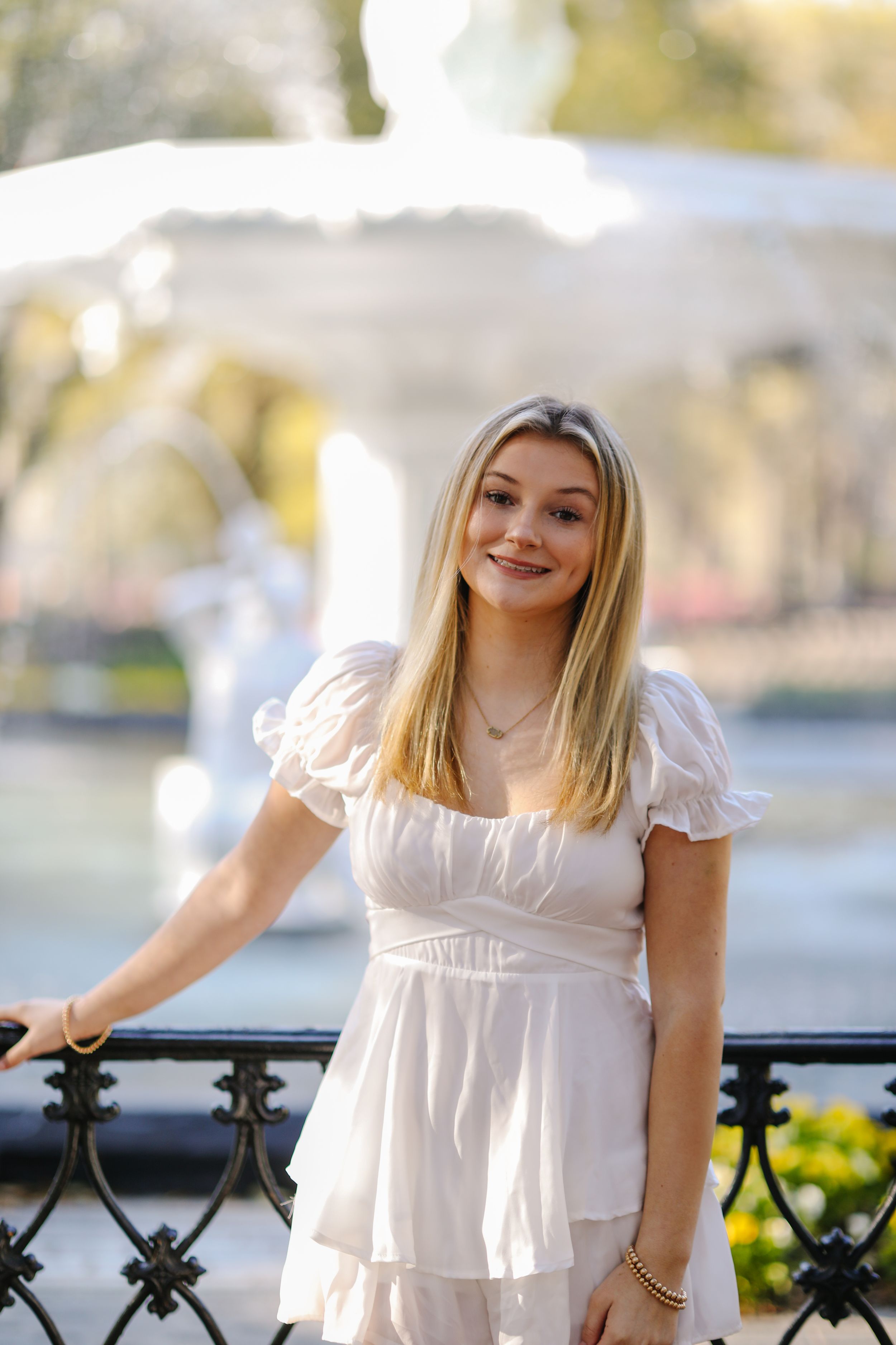 Young woman in white summer dress poses against ornate railing with blurred waterfront background in soft daylight.