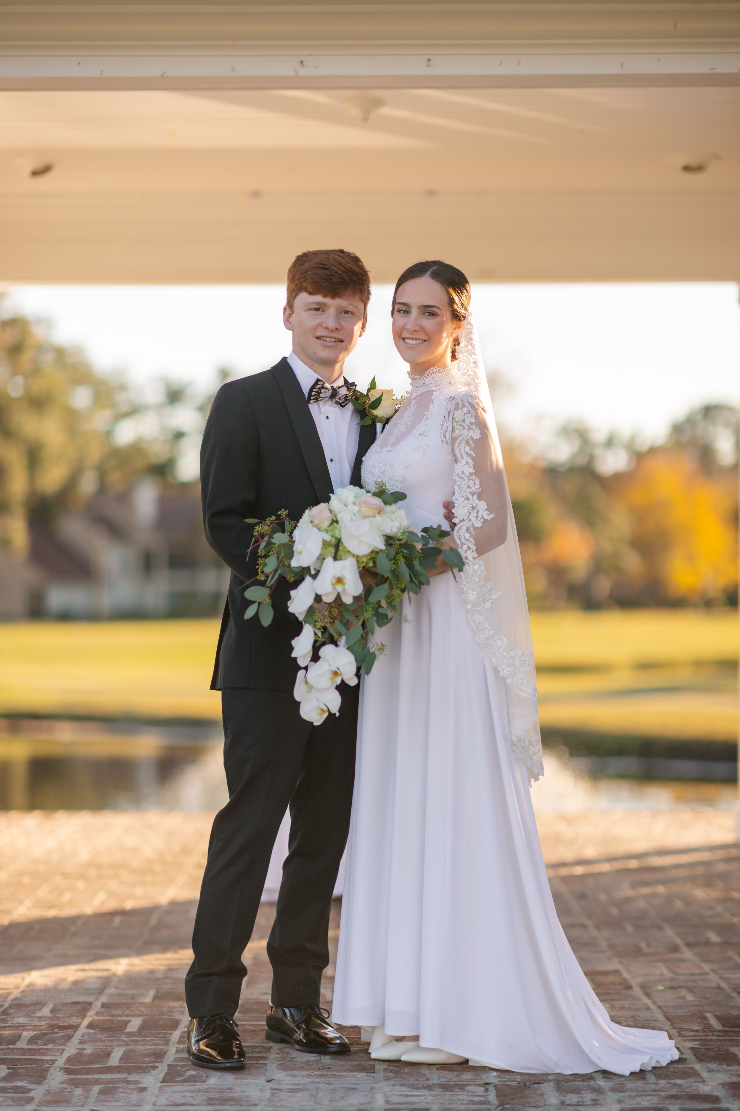 Bride and groom posing outdoors at golden hour, bride in lace gown holding white orchid bouquet, groom in black tuxedo.