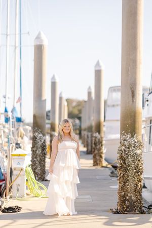 Blonde woman in white tiered dress posing on marina dock surrounded by wooden pylons and sailboats.