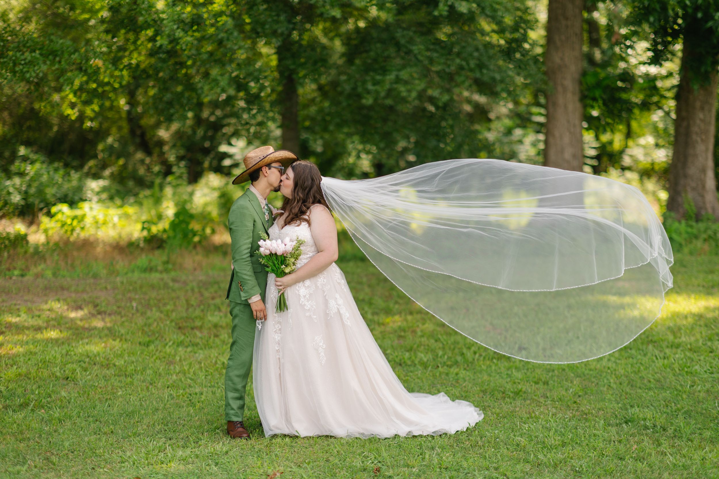 A bride and groom share a kiss outdoors as her long white veil flows dramatically in the breeze on their wedding day.
