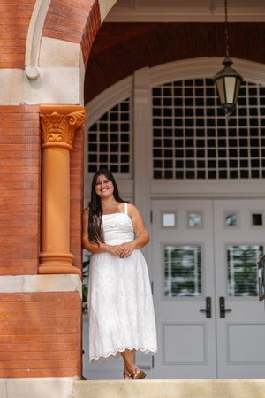 Young woman in white lace dress smiling beside ornate brick column and arched doorway of elegant building.