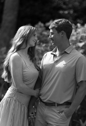Black and white photo of a young couple smiling at each other outdoors, woman in striped dress, man in polo shirt.
