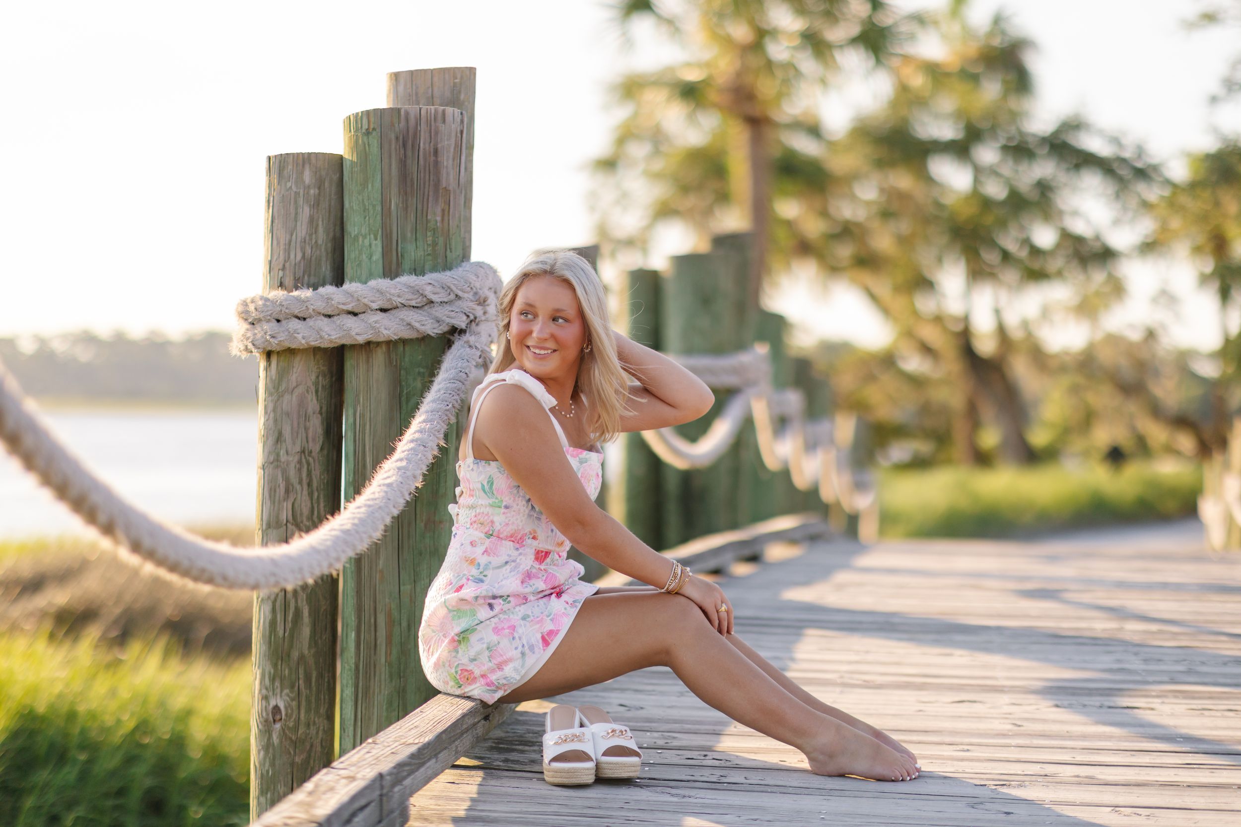 Smiling blonde woman in floral dress sitting on wooden boardwalk by water at golden hour.