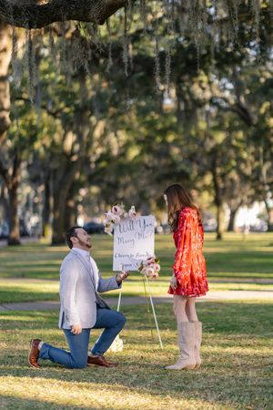Man on one knee proposing to woman in red dress outdoors under oak trees with a Will You Marry Me sign.
