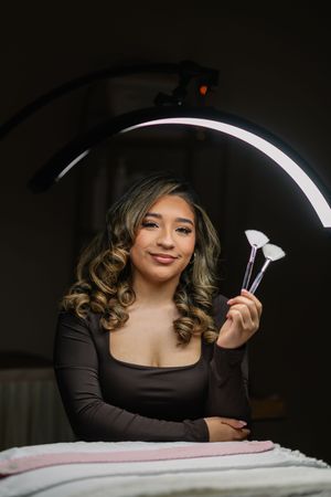 Young woman holding makeup brushes under a ring light, wearing a brown bodycon dress with curly highlighted hair.