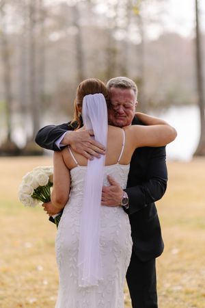 Emotional father hugging bride in white lace gown and veil, holding white roses bouquet, at outdoor wedding ceremony.