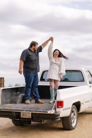 Couple dancing on the bed of a white pickup truck outdoors under a cloudy sky, woman in dress and teal boots.