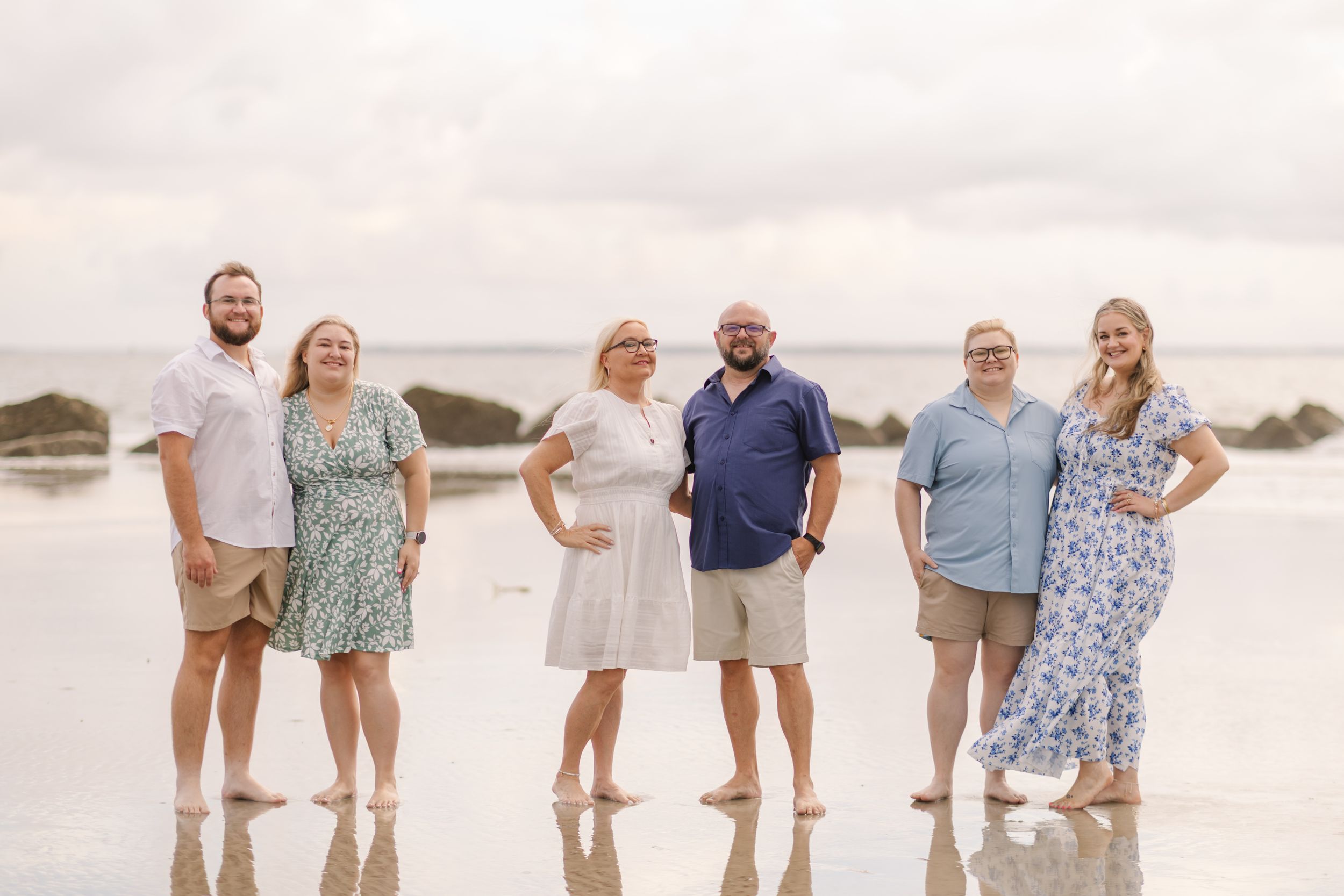 Six adults posing together on a reflective beach shore with cloudy skies in a casual family portrait.