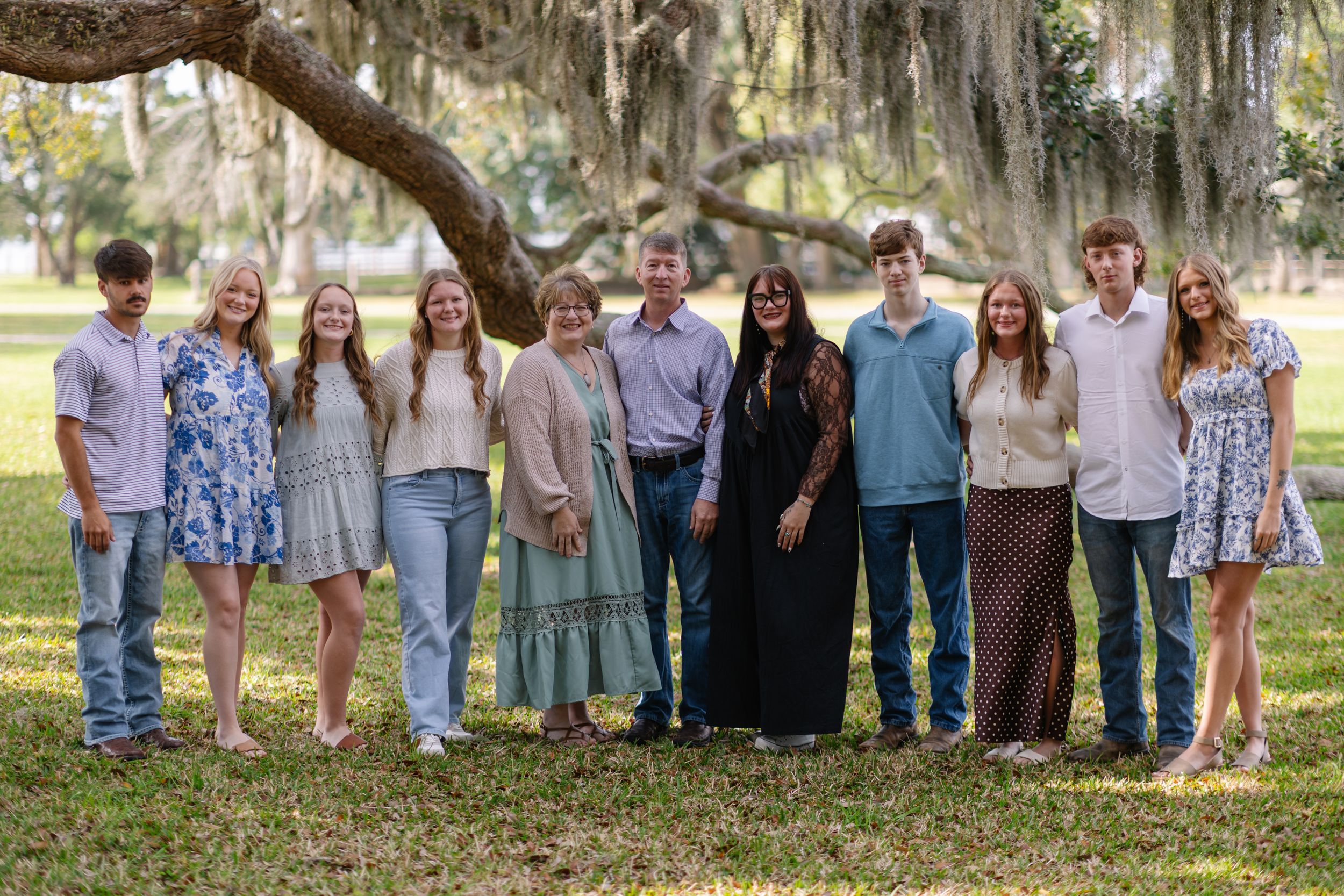 Large family group portrait of eleven people smiling outdoors under a sprawling oak tree on a sunny day.