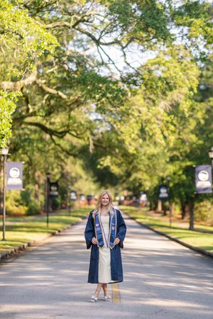 Young woman in navy graduation gown and stole smiling on a tree-lined campus walkway on a sunny day.