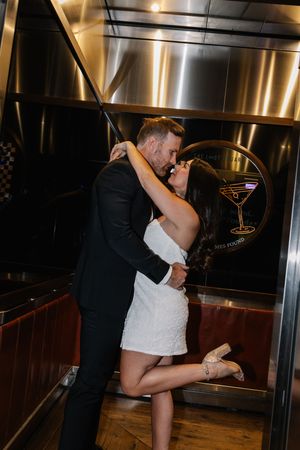 Couple sharing a romantic kiss in an elevator, woman in white dress lifting her leg, man in black suit.
