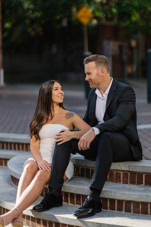 Couple sitting on brick steps smiling at each other, woman in white dress, man in black suit, urban outdoor setting.