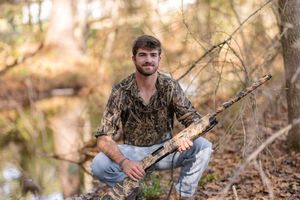 Young man in camo shirt crouching in forest with hunting rifle, surrounded by trees and natural woodland scenery.