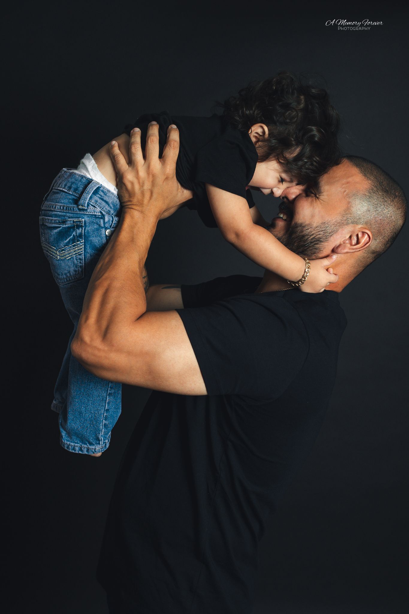 A muscular man in a black shirt lifts a curly-haired toddler close to his face against a dark background.