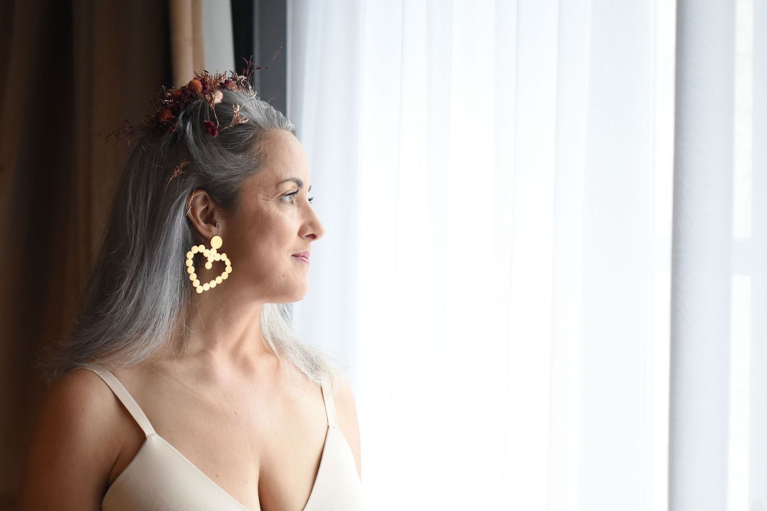 A woman with elegant gray hair and gold earrings gazes out a window in a contemplative pose.