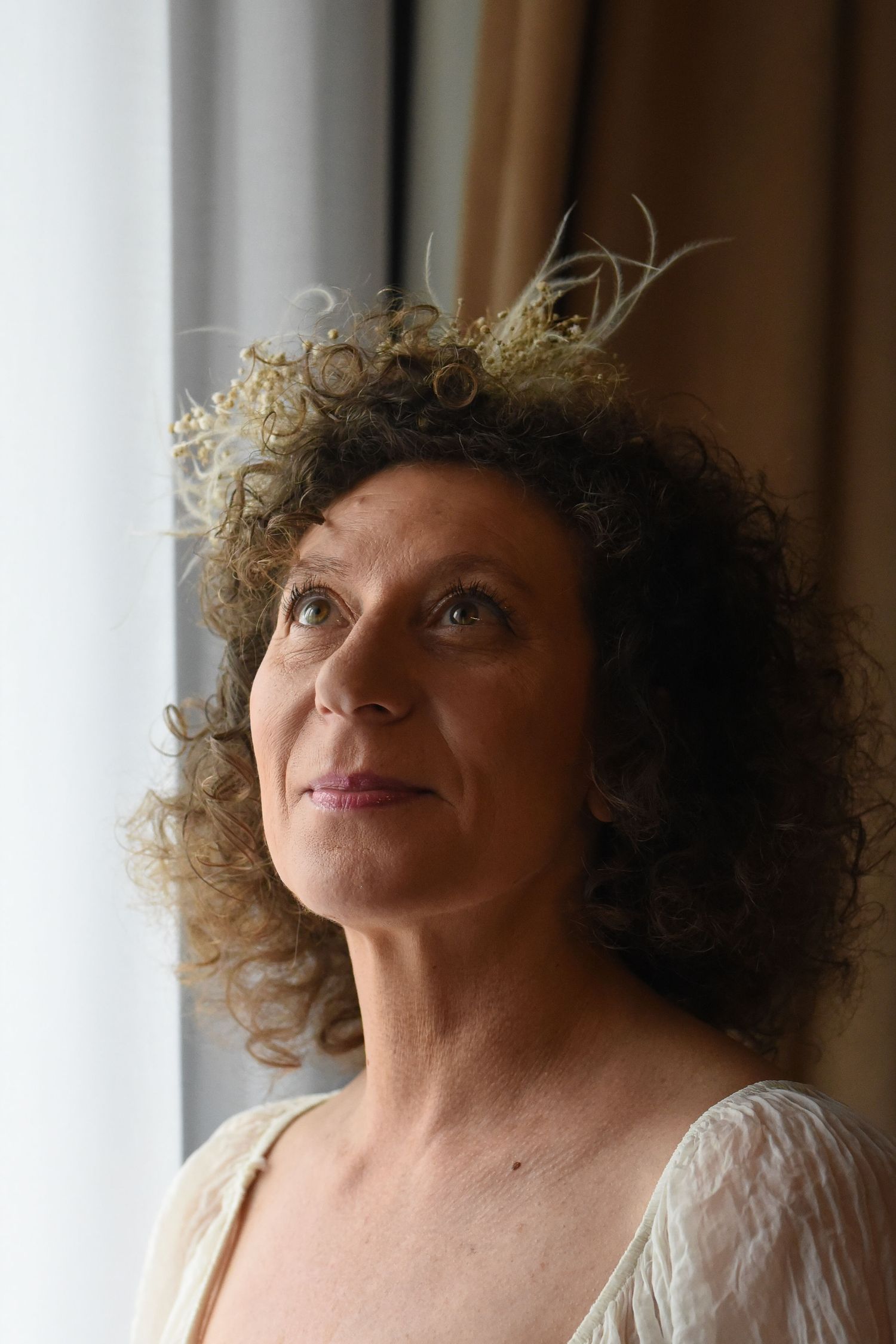 Person with curly dark hair wearing a white top gazes upward near a window in soft natural light.
