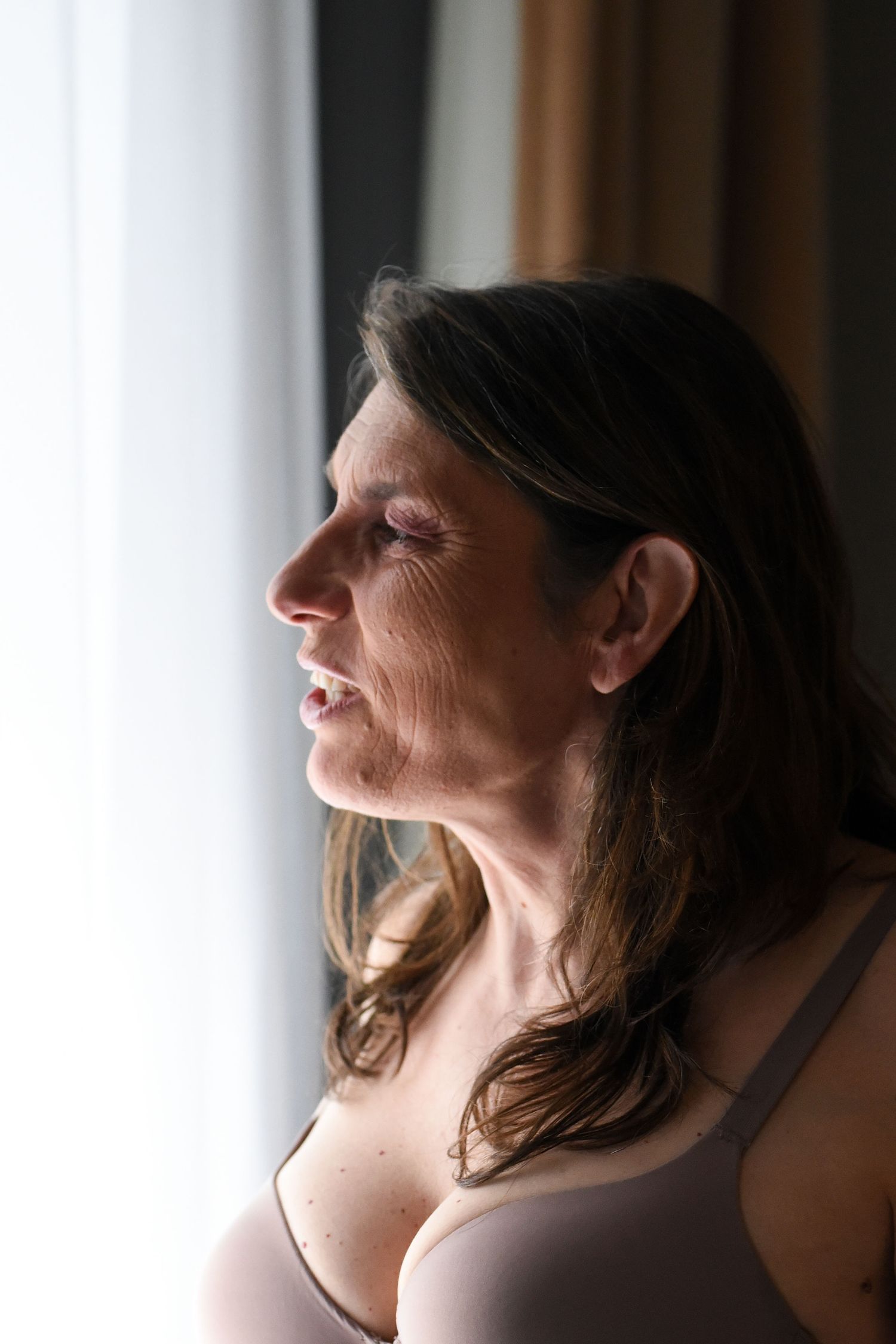 A profile view of a person in a beige bra smiling by a window with soft natural lighting.