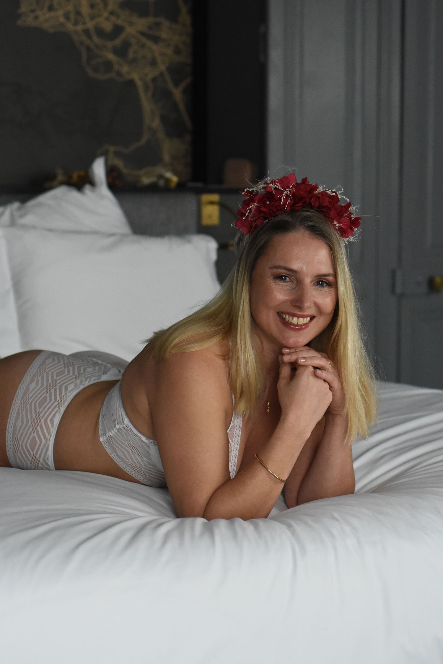 A person wearing a red flower crown lies on white bedding in a boudoir photography series.