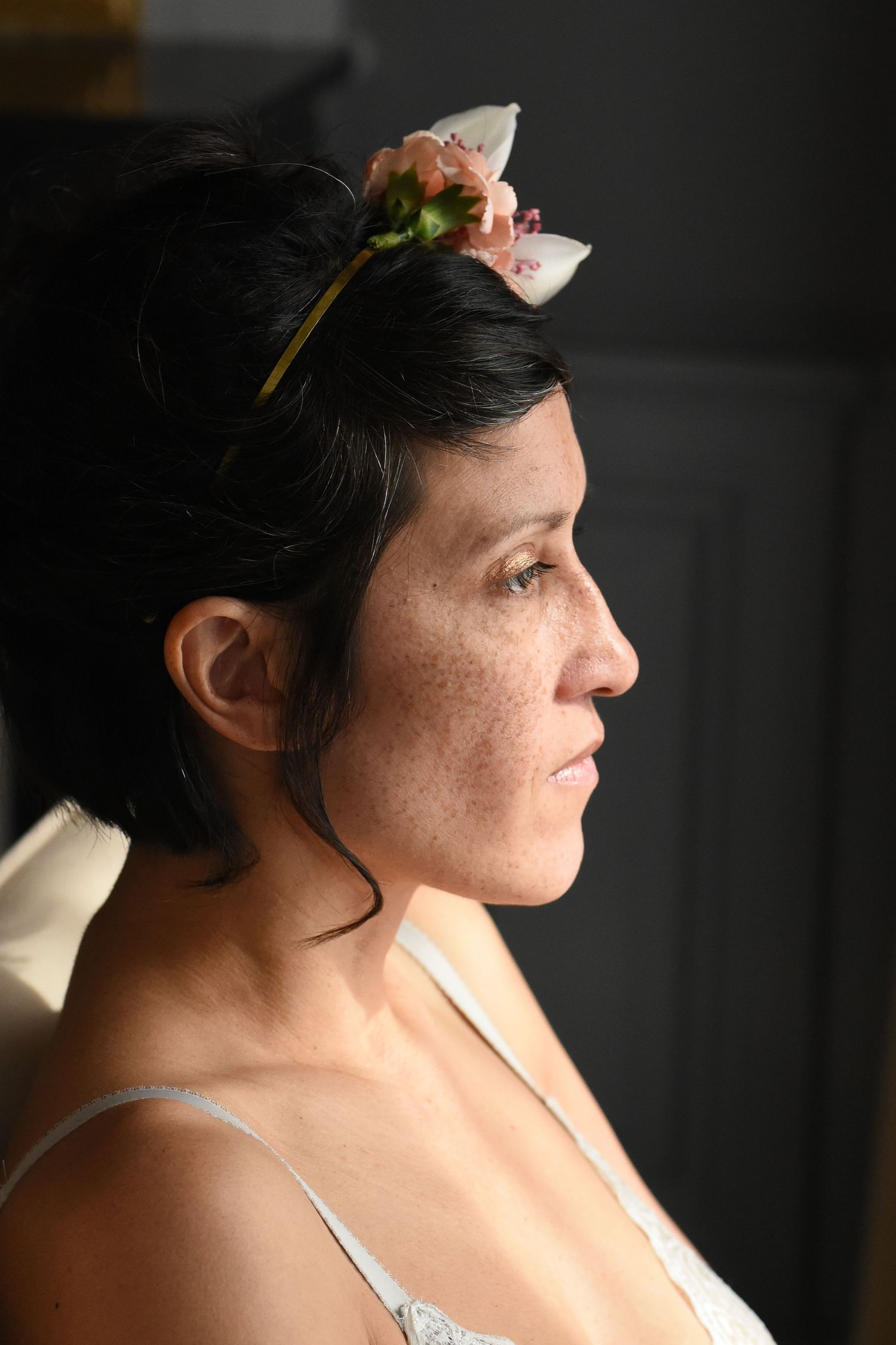 Side profile portrait with white floral accessory against dark backdrop.
