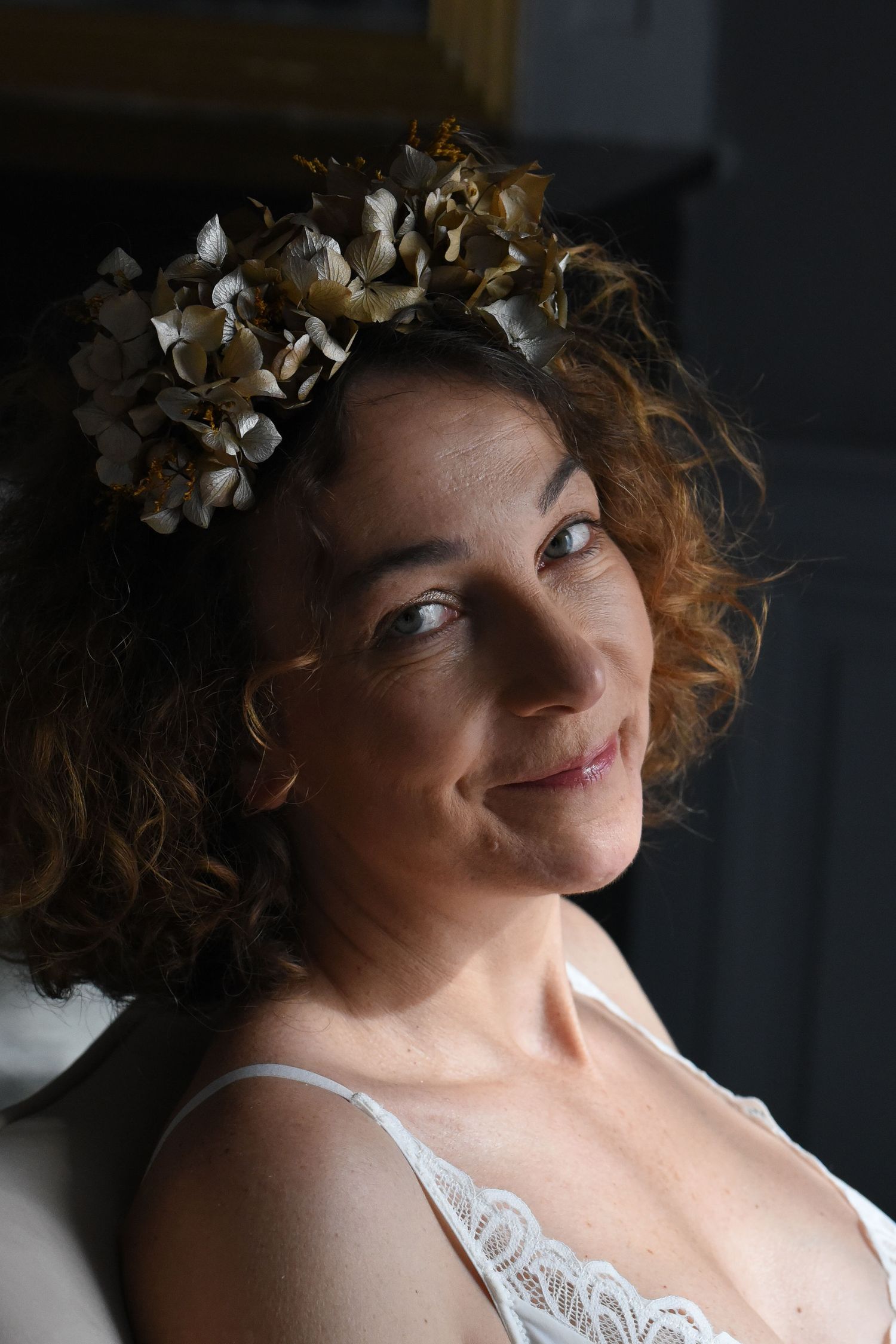 A portrait of someone with curly hair wearing a white lace top and floral crown in soft natural lighting.